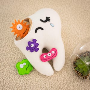 Può includere: Un dente di peluche bianco con una faccia triste, una lacrima e una cavità contenente un germe arancione. Il dente ha diversi germi colorati attaccati, tra cui viola, rosa e verde. Un terrario con una pianta succulenta è sullo sfondo.