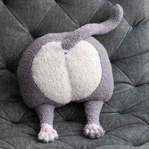 May include: A plush throw pillow shaped like the rear end of a cat, featuring gray and white textured sherpa fabric, a curved tail extending upward, and two gray legs with small pink paw pads visible at the bottom of the stuffed animal figure.