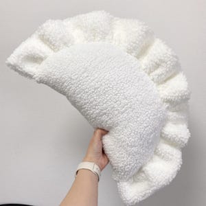 May include: A white, fluffy, dumpling-shaped pillow with a ruffled edge. The pillow is made of a soft, textured material, and is held by a person. The pillow is a decorative item.