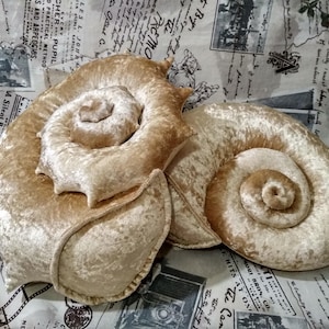 Puede incluir: Dos cojines de peluche con forma de conchas marinas en espiral, confeccionados en un suave tejido de terciopelo beige claro. Cada cojín presenta un diseño de concha enrollada con una superficie texturizada y brillante y bordes redondeados.