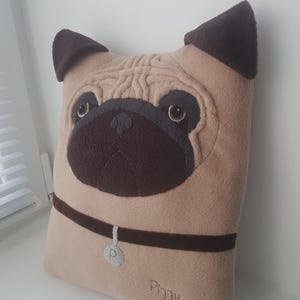 Puede incluir: Un coj&iacute;n beige con forma de pug con detalles marrones. El coj&iacute;n presenta una cara bordada detallada, incluyendo ojos, nariz y orejas. Un collar marr&oacute;n con una etiqueta plateada y el nombre "Piggy" bordado.