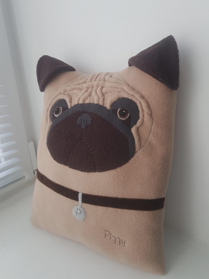Puede incluir: Un coj&iacute;n beige con forma de pug con detalles marrones. El coj&iacute;n presenta una cara bordada detallada, incluyendo ojos, nariz y orejas. Un collar marr&oacute;n con una etiqueta plateada y el nombre "Piggy" bordado.
