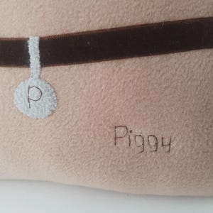 Puede incluir: Una cama beige para mascotas con un collar de terciopelo marr&oacute;n. Una etiqueta blanca con la letra "P" est&aacute; unida al collar. La palabra "Piggy" est&aacute; bordada con hilo marr&oacute;n en la cama.