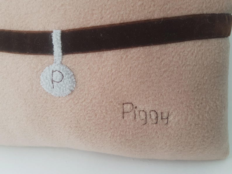 Puede incluir: Una cama beige para mascotas con un collar de terciopelo marr&oacute;n. Una etiqueta blanca con la letra "P" est&aacute; unida al collar. La palabra "Piggy" est&aacute; bordada con hilo marr&oacute;n en la cama.