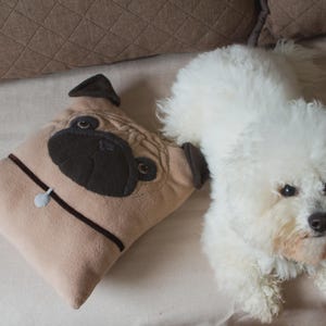 Puede incluir: Un coj&iacute;n beige con forma de pug con detalles negros y marrones descansa junto a un perro blanco y esponjoso. El coj&iacute;n tiene un collar marr&oacute;n oscuro y una peque&ntilde;a etiqueta azul claro. El perro tiene un hocico corto y ojos oscuros.