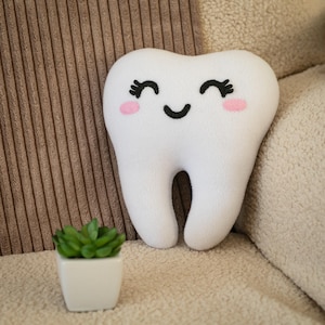 May include: A white plush tooth-shaped pillow with a smiling face, black eyelashes, and pink cheeks. The pillow is on a beige surface with a small potted succulent plant in a white square pot.