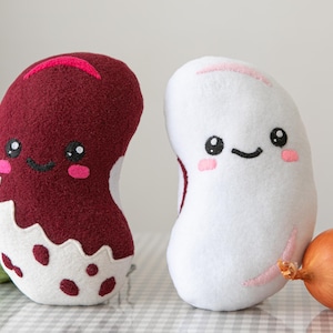 以下が含まれることがあります： 笑顔の顔をした、豆の形をした2つのぬいぐるみ。一つは白のアクセントが付いたバーガンディ色、もう一つはピンクのアクセントが付いた白です。キャベツと玉ねぎも画像にあります。
