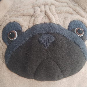 Puede incluir: Primer plano de un coj&iacute;n con la cara de un pug beige. La cara del pug presenta un hocico gris oscuro, detalles de ojos azul-gris y ojos marrones. El coj&iacute;n tiene un aspecto texturizado y afelpado.