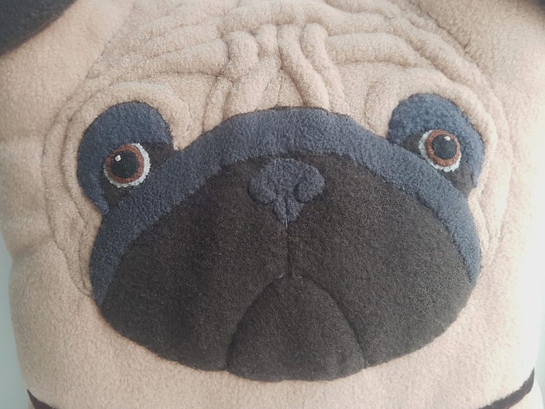 Puede incluir: Primer plano de un coj&iacute;n con la cara de un pug beige. La cara del pug presenta un hocico gris oscuro, detalles de ojos azul-gris y ojos marrones. El coj&iacute;n tiene un aspecto texturizado y afelpado.