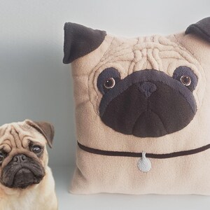 Puede incluir: Un coj&iacute;n beige con forma de pug con detalles en marr&oacute;n oscuro, incluyendo las orejas, la nariz y el collar. El coj&iacute;n tiene un dise&ntilde;o texturizado y una peque&ntilde;a etiqueta redonda en el collar. Un perro pug real est&aacute; al lado del coj&iacute;n.