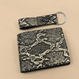 May include: A black and gold snakeskin-patterned wallet and matching keyring. The wallet is folded and rectangular. The keyring has a silver ring. The items are displayed on a neutral background.