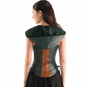 May include: A dark green leather corset featuring a hood, a central vertical brown leather panel, and side lacing with thin black cords. The garment is sleeveless and has a fitted silhouette that extends to the waist, worn over a black skirt.