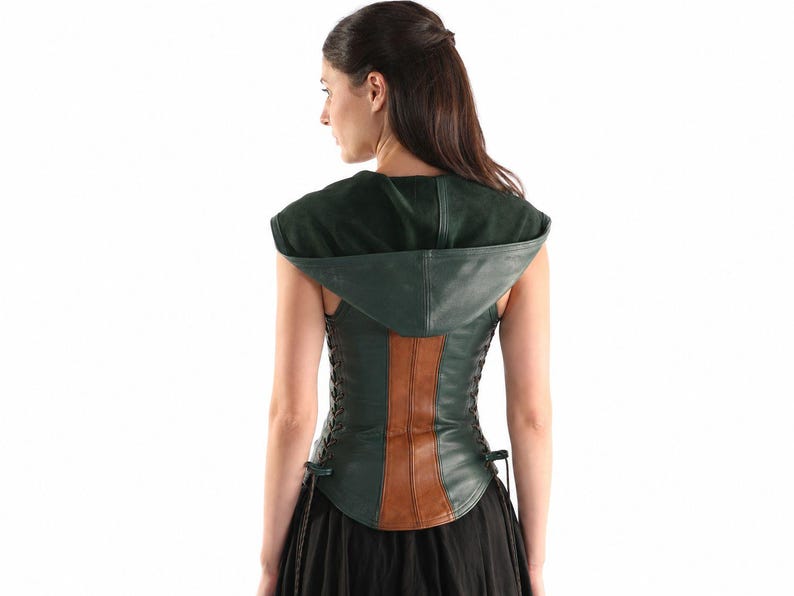 May include: A dark green leather corset featuring a hood, a central vertical brown leather panel, and side lacing with thin black cords. The garment is sleeveless and has a fitted silhouette that extends to the waist, worn over a black skirt.