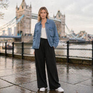 Puede incluir: Una persona con una chaqueta vaquera azul, una camiseta blanca y pantalones negros de pernera ancha está de pie frente al Tower Bridge en Londres. También lleva zapatillas blancas. El fondo es un día nublado.