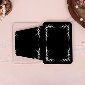 May include: A black rectangular card holder featuring a white thorn or barbed wire pattern printed along the edges. The holder is made of a clear plastic material and has a flap closure that secures the contents within the central compartment.