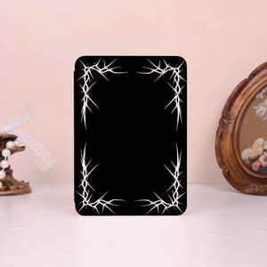 May include: A rectangular black tablet case featuring a white graphic design of thorny vines or sharp, pointed branches framing the four corners of the front cover. The design consists of thin, intersecting lines that create a sharp, angular pattern.
