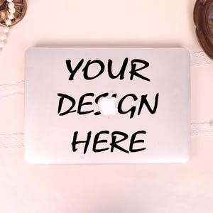 May include: A silver laptop with the words "YOUR DESIGN HERE" in black lettering. The laptop has a white Apple logo in the center. The background is a light pink color with decorative elements.