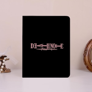 May include: A rectangular black notebook featuring the text DEATH NOTE in a light pink, distressed serif font centered on the front cover, with Japanese characters positioned directly below the English title in a matching light pink color.