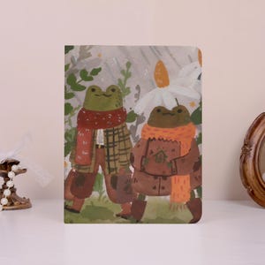 May include: A decorative notebook featuring an illustration of two anthropomorphic frogs. One frog wears a plaid jacket and red scarf, the other a brown coat and orange scarf. The background includes flowers and foliage.