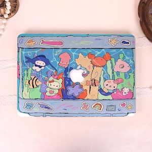 May include: A blue laptop case featuring a colorful underwater scene with cartoon fish, a crab, and other sea creatures. The case has a white Apple logo and a light blue border. The design is whimsical and playful.