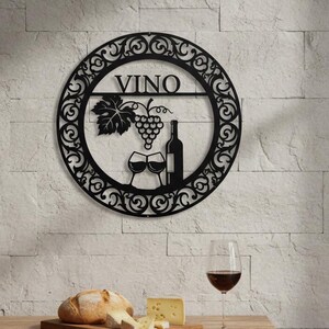 Puede incluir: Arte de pared de metal negro con la palabra "VINO" e imágenes relacionadas con el vino, incluyendo un racimo de uvas, una botella y copas. El arte está montado en una pared de ladrillo blanco texturizado. Una mesa de madera exhibe pan, queso y una copa de vino tinto.