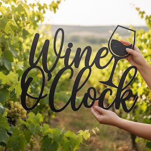 Op de afbeelding: Een wandbord van zwart metaal met de cursieve tekst Wine O'Clock en een silhouet van een wijnglas. Het bord wordt vastgehouden tegen een achtergrond van groene wijnranken in een wijngaard, wat het uitgesneden ontwerp van de letters en het glas toont.