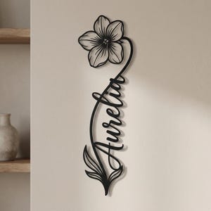 Könnte beinhalten: Ein schwarzes Wandkunstobjekt aus Metall, das oben eine einzelne Blume zeigt, deren langer, geschwungener Stiel den Namen Aurelia in Schreibschrift bildet und unten in zwei spitzen Blättern endet, montiert an einer hellen, einfarbigen Wand.