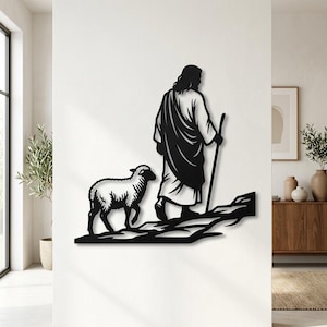 Puede incluir: Una pieza de arte mural de metal negro que representa a una figura con túnica caminando con un bastón, acompañada por una oveja en un camino rocoso. El diseño de silueta presenta líneas limpias y un acabado plano, mostrando a la figura de espaldas.