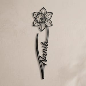 Könnte beinhalten: Ein Wandkunstobjekt aus schwarzem Metall in Form einer einzelnen Narzissenblüte. Der Stängel der Blume ist mit dem in Schreibschrift geschriebenen Namen Nanibe verbunden, der vertikal entlang des Stängels verläuft und ein durchgehendes Design bildet.