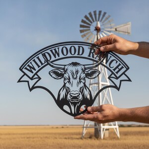 Könnte beinhalten: Ein schwarzes Metallschild mit einer detaillierten Illustration eines Kuhkopfes unter einem geschwungenen Banner mit dem Text WILDWOOD RANCH. Das Design ist aus einem einzigen Stück Metall mit klaren Linien und einer matten Oberfläche gefertigt.