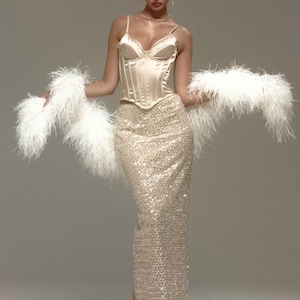 May include: A champagne-coloured corset top with spaghetti straps and a sequined, floor-length skirt. The outfit is accessorized with a white feather boa. The overall look is elegant and suitable for a special occasion.