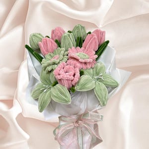 May include: A fabric flower bouquet featuring pink and green blooms, wrapped in white paper and tied with a pink ribbon. The arrangement includes tulips, lilies, and other flower varieties. Set against a cream-coloured fabric backdrop.