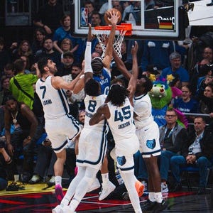 Puede incluir: Un partido de baloncesto en curso, con jugadores con uniformes blancos y azules saltando para alcanzar el balón cerca del aro. Los jugadores llevan zapatillas deportivas. La multitud es visible en el fondo.