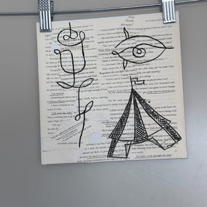 Könnte beinhalten: Ein quadratisches Blatt Papier mit gedrucktem Buchtext und einer schwarzen Tuschezeichnung einer Rose und des Eiffelturms. Die Zeichnung enthält eine stilisierte Augenform über dem Turm. Das Papier wird von zwei silbernen Metallklammern gehalten.