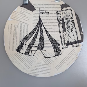 Könnte beinhalten: Eine kreisförmige Collage aus Buchseiten mit einer schwarzen Tuschezeichnung eines Zirkuszeltes und einer Eintrittskarte. Der Text auf dem Ticket lautet "Admit one", "Come one Come all", "Amazing show" und "Circus 16-24 July". Das Kunstwerk wird von zwei Metallklammern gehalten.