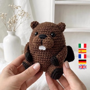May include: A handmade crochet beaver plushie in shades of brown and black. The beaver has white teeth, black eyes, and a black nose. The plushie is held in a person's hands.