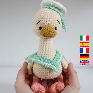 Puede incluir: Muñeca de pato de ganchillo hecha a mano con cuerpo color crema, pico amarillo y ojos negros. El pato lleva un gorro y bufanda de marinero verde claro y blanco, y tiene pies rosas. La muñeca está sostenida en manos.