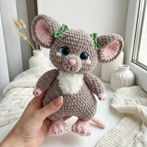 Puede incluir: Un juguete de ratón de ganchillo hecho a mano en tonos grises, rosas y blancos. El ratón tiene orejas grandes con el interior rosa, ojos azules y una nariz rosa. Tiene pequeños lazos verdes en las orejas y un pecho blanco y esponjoso.