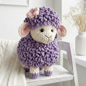 Puede incluir: Un juguete de oveja de peluche hecho a mano en tonos de morado, crema y rosa. La oveja tiene un cuerpo suave y texturizado, una cara color crema y orejas forradas de rosa. Una flor rosa adorna su cabeza. El juguete está sobre una silla de madera blanca.