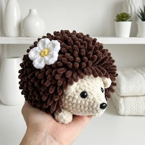 May include: A crocheted hedgehog toy in brown and cream yarn, with a white flower and yellow centre. The toy has black safety eyes and is held in a hand. The toy is a soft, plush design.