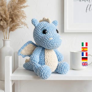 May include: A light blue crocheted dragon plush toy with cream-colored accents on its belly, wings, and head. The dragon has large, black eyes and is sitting on a white shelf. A white mug is to the right of the dragon.