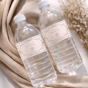 May include: Two clear plastic water bottles with white caps, each featuring a beige and white gingham patterned label. The label displays a decorative frame with a bow, the text A BABY IS brewing, a coffee cup icon, and OLIVIA'S BABY SHOWER.