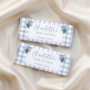 Könnte beinhalten: Zwei rechteckige Schokoriegelverpackungen mit hellblau-weißem Vichy-Muster. Das weiße Etikett in der Mitte zeigt den Text Charlotte's BABY SHOWER SATURDAY, 18.09.XX, verziert mit Aquarellillustrationen von Blaubeeren und blauen Schleifen.