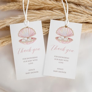 Könnte beinhalten: Zwei weiße Babyparty-Geschenkanhänger mit einer Aquarellillustration einer rosa Muschel mit Perlen. Der Text "Thank you" ist in Schreibschrift geschrieben, mit dem Text "Für das Verwöhnen unseres Babys mit Liebe" und "Anna's Baby Shower".