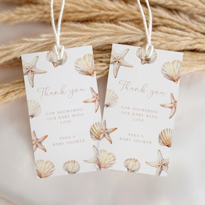 Op de afbeelding: Twee rechthoekige baby shower cadeaulabels met een patroon van schelpen en zeesterren. De labels lezen "Bedankt" en "Voor het verwennen van onze baby met liefde, Anna's Baby Shower."
