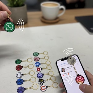 Op de afbeelding: Sleutelhangers met social media logo's, waaronder Instagram, WhatsApp en YouTube. Elke sleutelhanger heeft een gouden ketting en ring. Eén sleutelhanger is verbonden met een smartphone, met een signaalafbeelding. Een kopje koffie en een plant staan op de achtergrond.