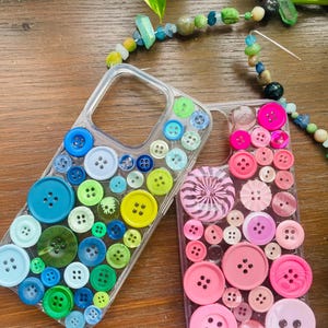 May include: Two clear phone cases decorated with colorful buttons. One case features blue, green, and yellow buttons. The other case is adorned with pink buttons and a swirl design. A beaded necklace is visible in the background.