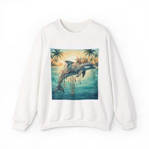 Puede incluir: Sudadera blanca con una ilustración en acuarela de un delfín adornado con flores y atrapasueños. El delfín está situado frente a una escena de playa tropical con palmeras y una puesta de sol. El diseño está centrado en la parte delantera.