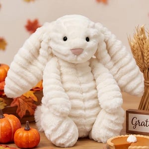 Puede incluir: Un conejo de peluche color crema con orejas largas y caídas y una nariz rosada. El peluche está sentado erguido, rodeado de calabazas, hojas de otoño y un pastel. El conejo tiene un aspecto texturizado, similar a la pana.