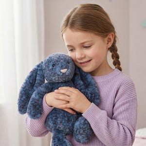 Puede incluir: Un peluche azul con forma de conejo, con largas orejas y una nariz rosa, es sostenido por una persona que lleva un suéter morado claro. El conejo tiene un aspecto suave y texturizado. La persona sonríe y parece abrazar el peluche.